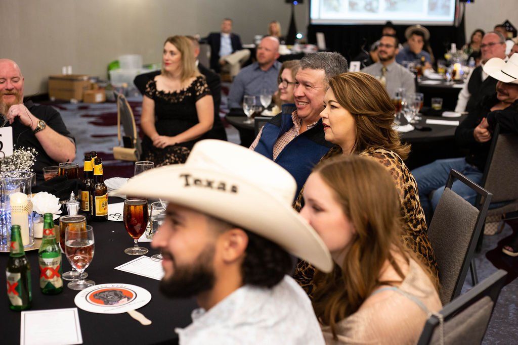 People seated at tables during an indoor event, focused on a presentation shown on a screen in the background.