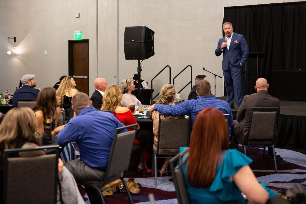 A man in a suit speaks into a microphone before an audience seated at tables in a conference room.