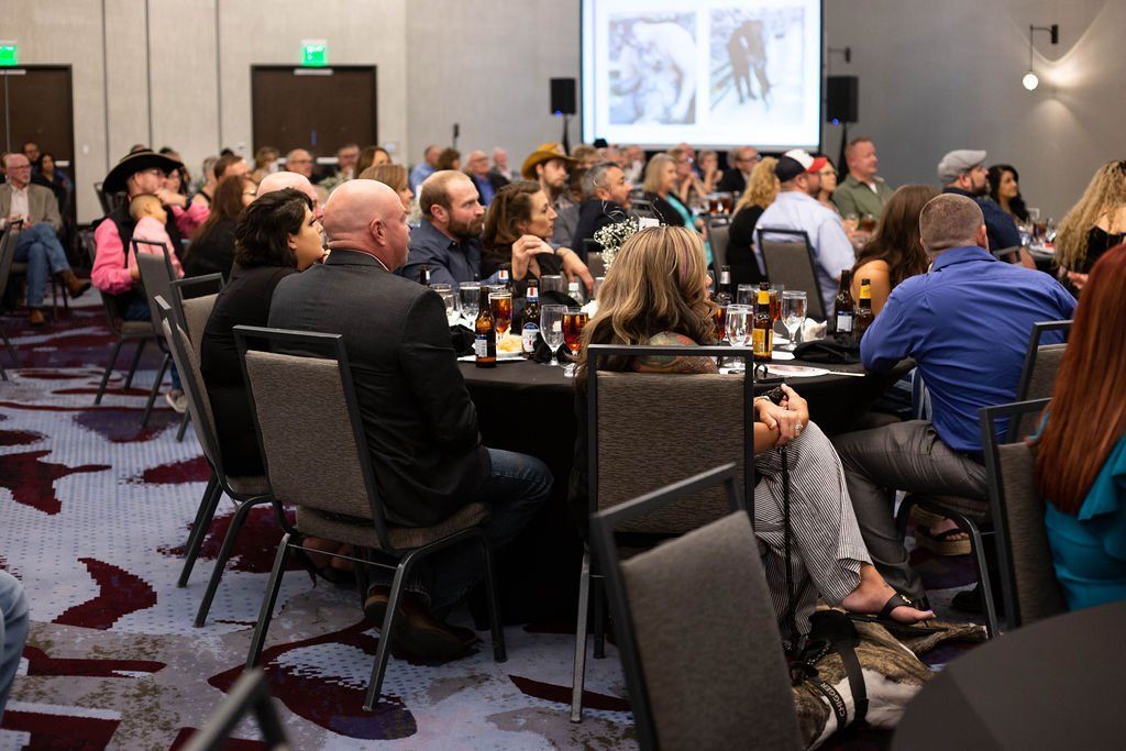 Attendees sit at round tables in a conference hall facing a presentation screen, with some guests dining.