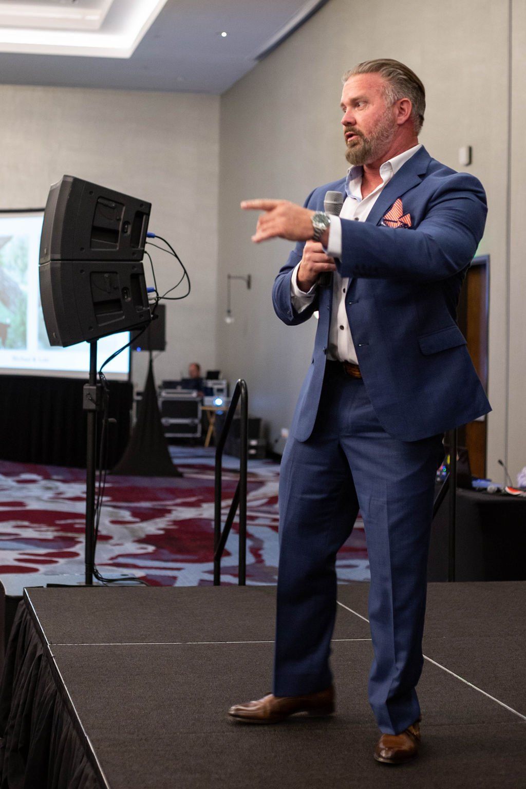 A presenter in a blue suit stands on a stage holding a microphone and gesturing to the side during a conference.