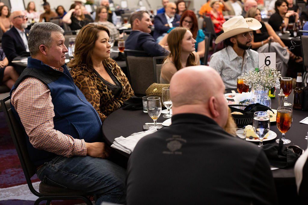A group of people sit at round tables in a conference hall, listening to a presentation during an event.