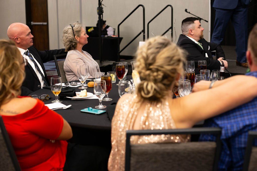 Guests at a banquet table sit in a dim room with black tablecloths, watching an event.