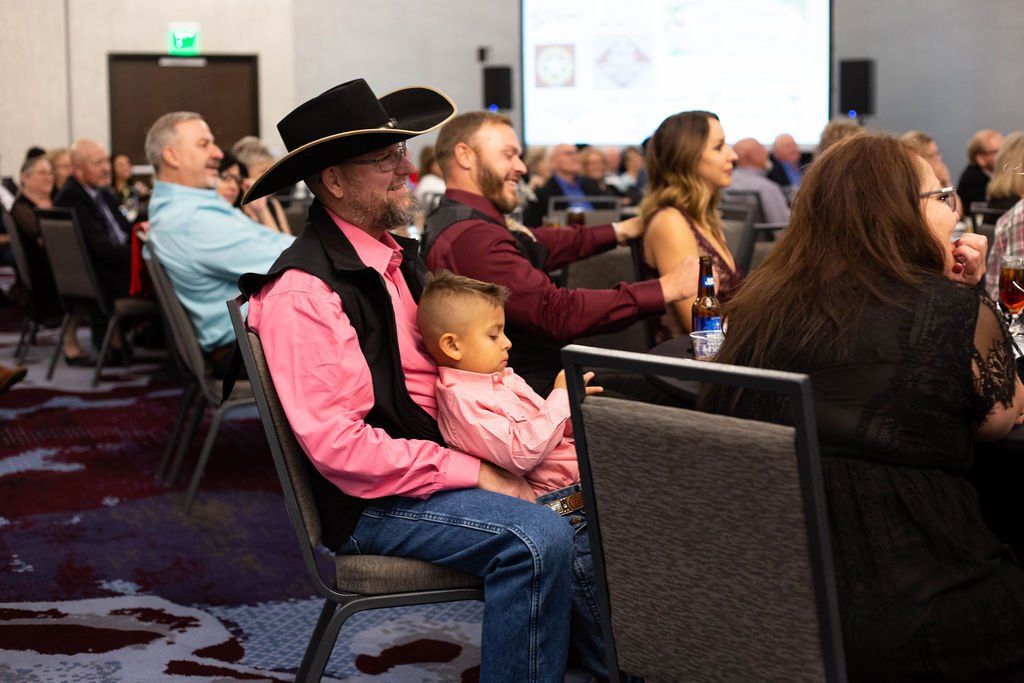 A man in a black hat holds a young child on his lap in a crowded, dimly lit event hall with people seated at tables.
