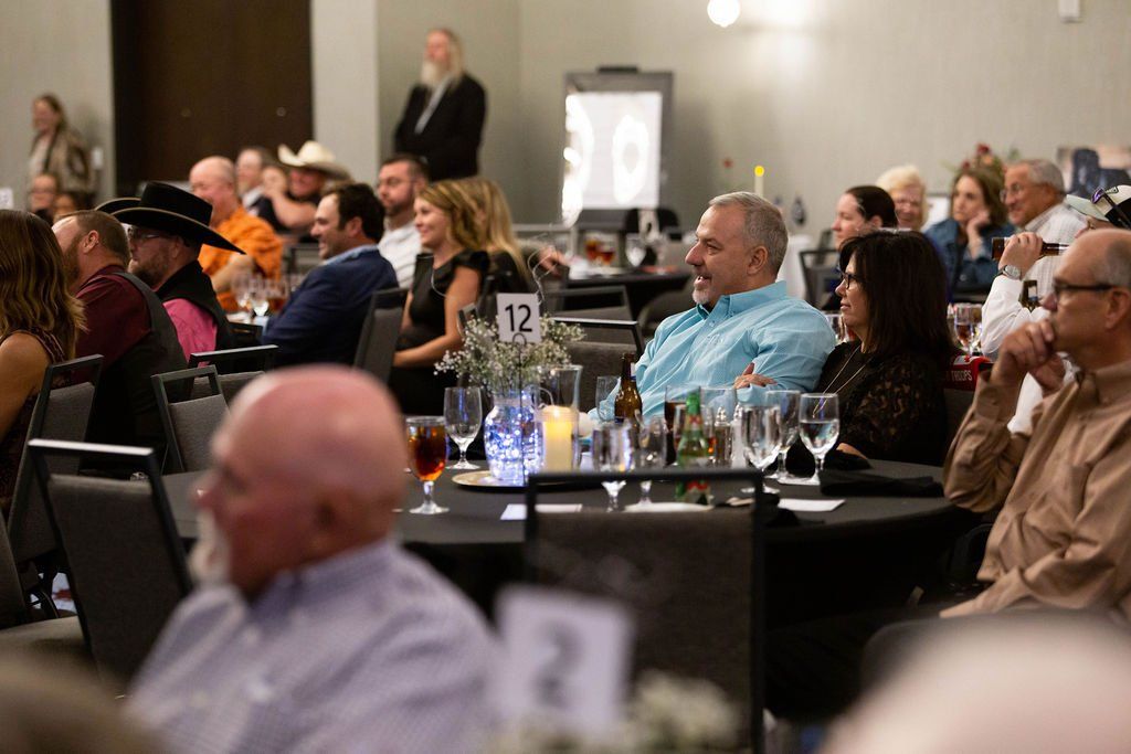 People sit at round tables in a dimly lit conference room, listening to a presentation or speech.