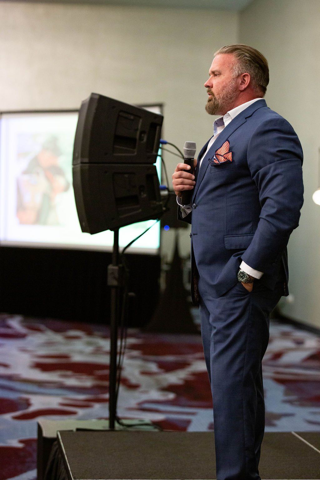 A person in a blue suit speaking into a microphone on a stage in front of a projector screen.
