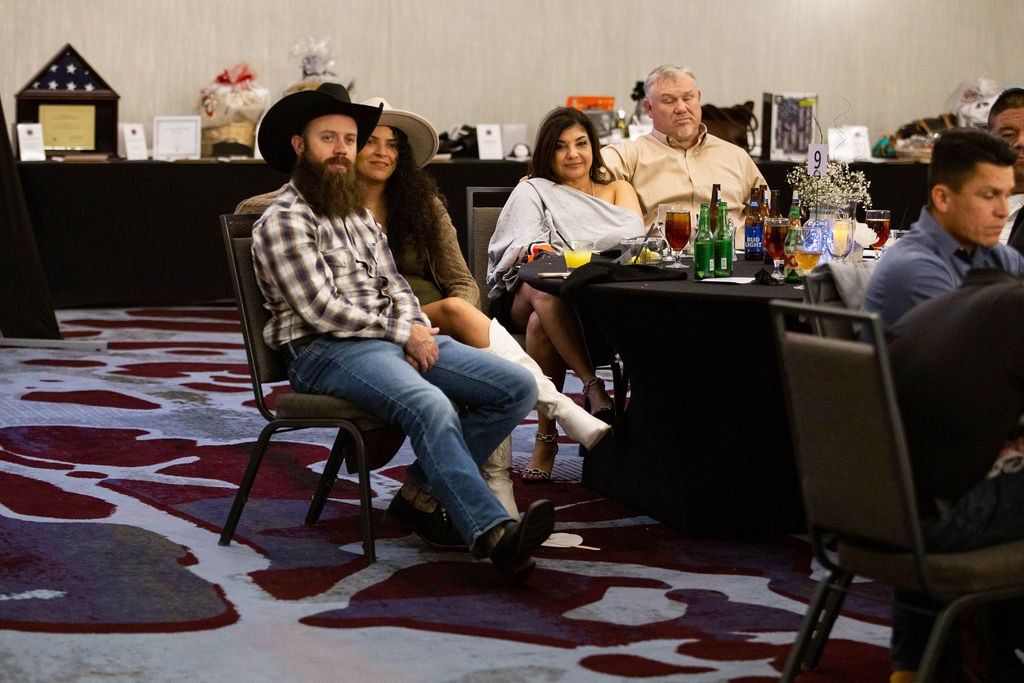 People seated at tables in a ballroom with patterned carpet, attending an event with drinks and gift baskets.