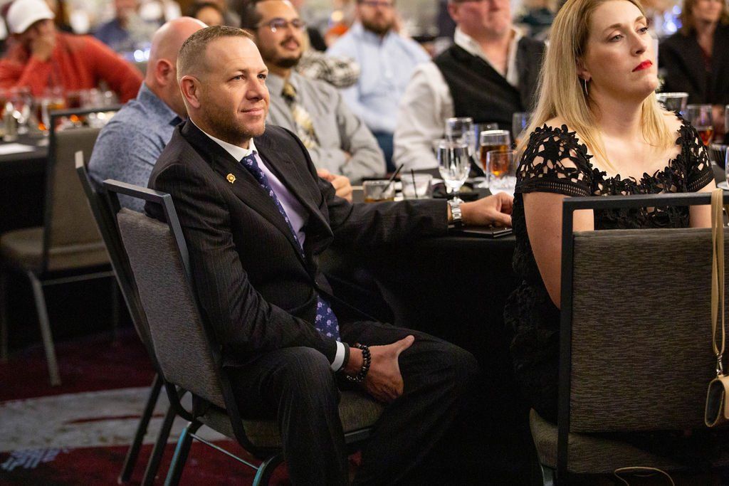 People seated at tables in a banquet hall, with a person in a suit and tie looking toward the front of the room.
