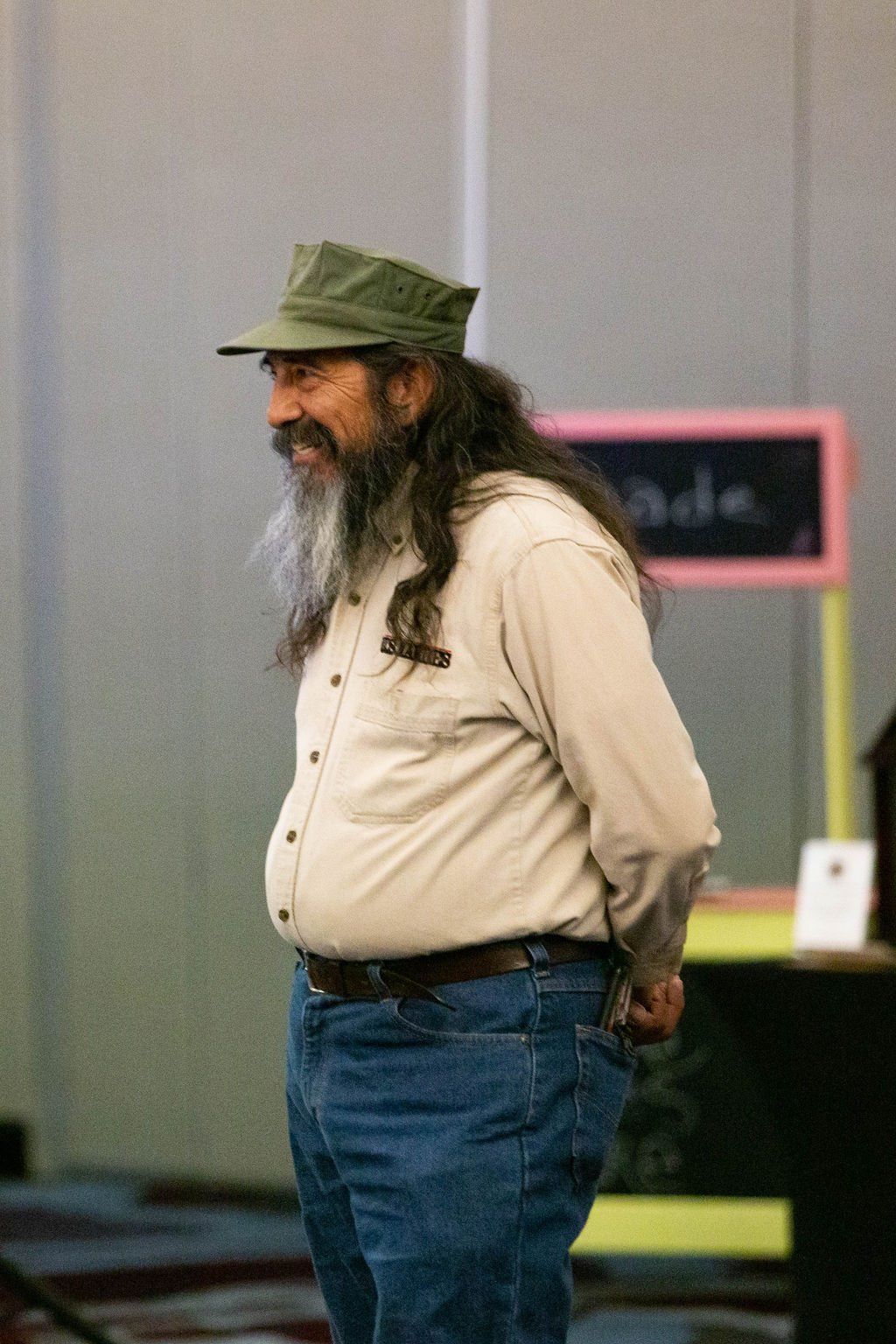 A smiling person with a long beard and green cap wears a tan shirt and jeans, standing indoors by a small chalkboard.