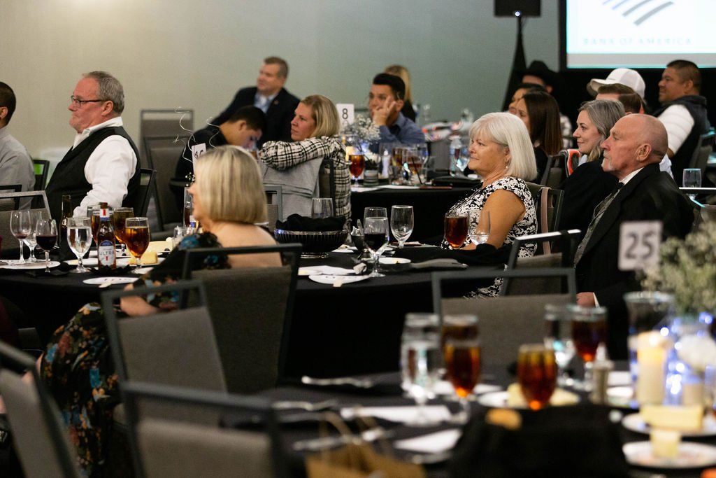 People in formal wear seated at round tables during a banquet or presentation in a well-lit event space.