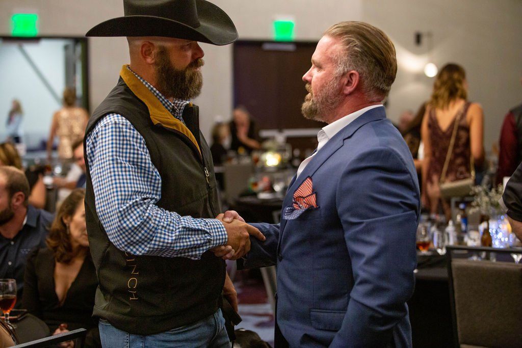 Two men shaking hands at an indoor event; one wears a cowboy hat and vest, the other a blue suit jacket.