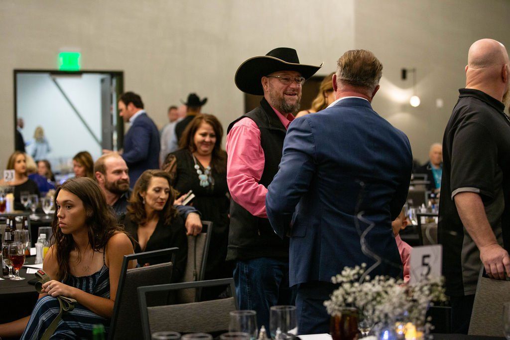 People chat at a formal indoor event, with a man in a cowboy hat and a woman in a blue striped dress in the foreground.