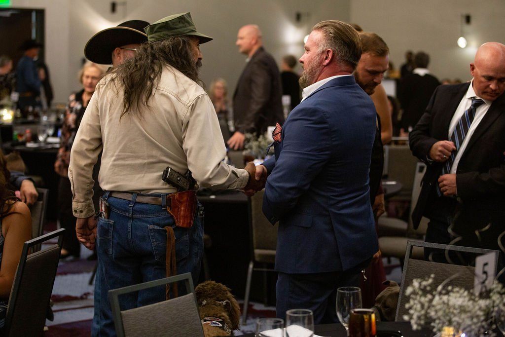 Two individuals, one in casual Western attire and one in a formal suit, shaking hands in a crowded event hall.