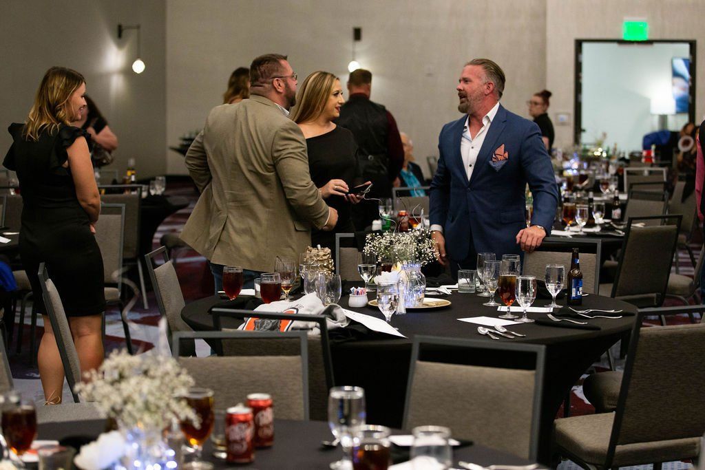 People in formal attire gather around tables for an event in a dimly lit banquet hall.