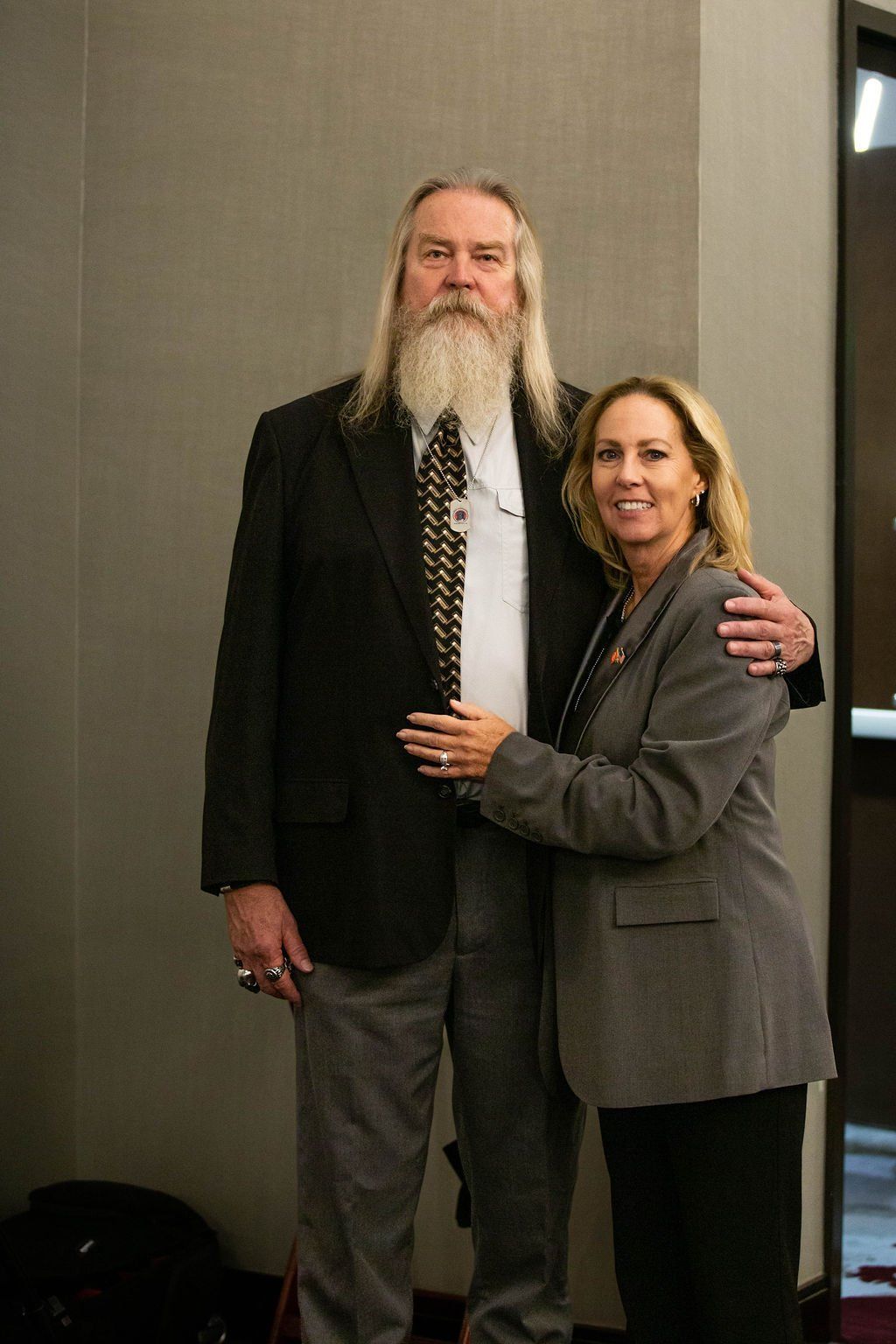 A smiling person in a gray blazer embraces a tall, bearded man in a suit and patterned tie indoors.
