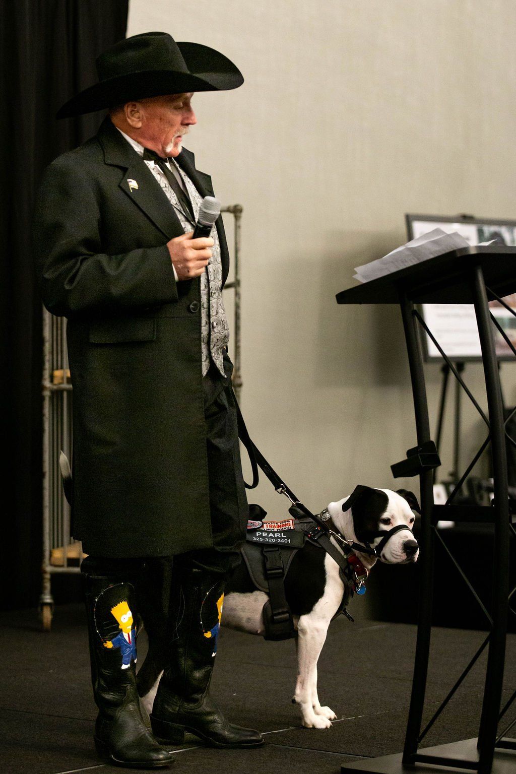 A person in a black coat, hat, and boots stands at a podium holding a microphone, with a service dog on a leash nearby.