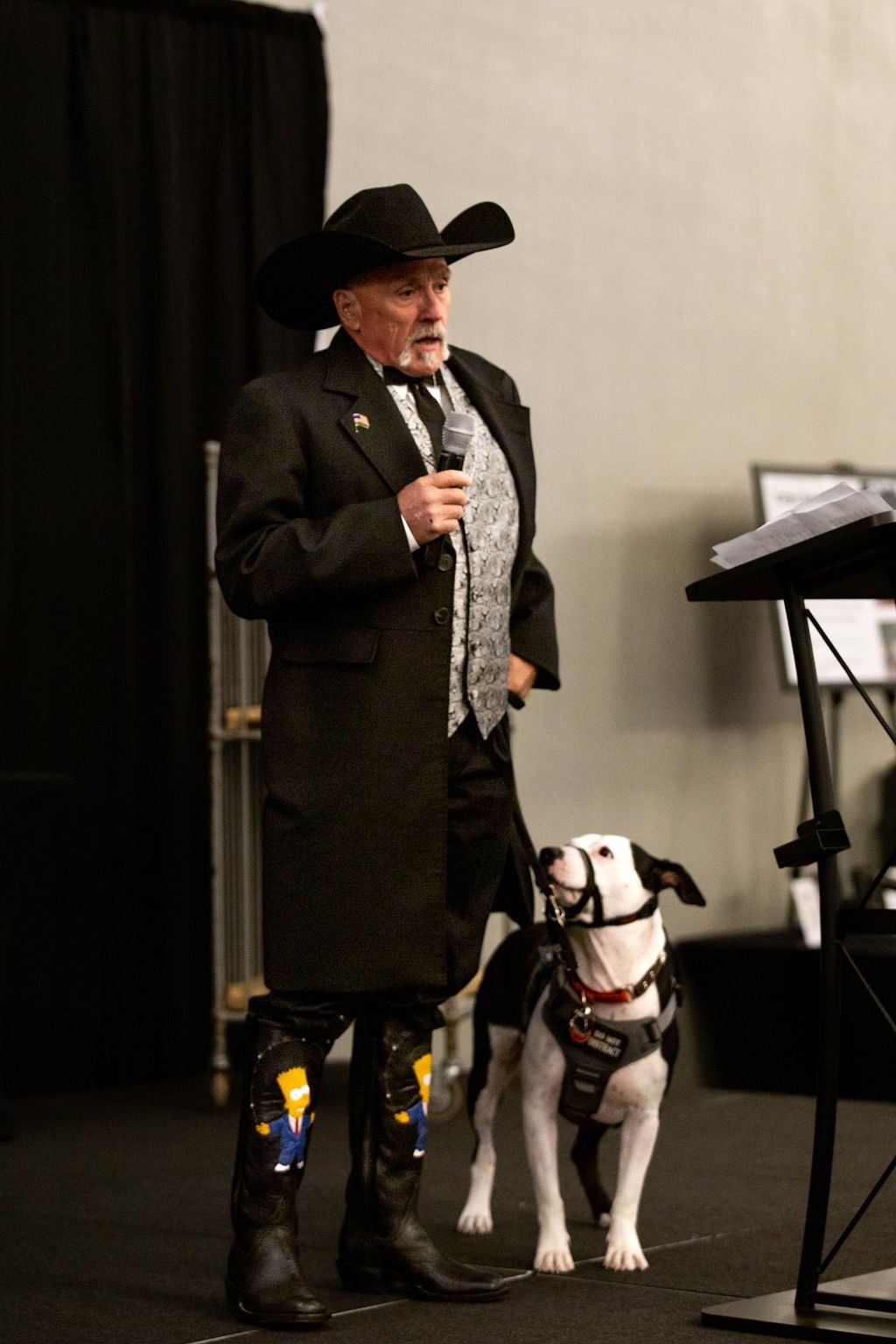 A person in a cowboy hat and suit speaks at a podium with a service dog standing by their side.