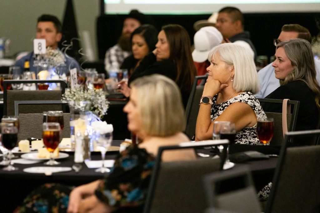 Attendees at a formal event sit at round tables, listening to a presentation in a dimly lit conference room.