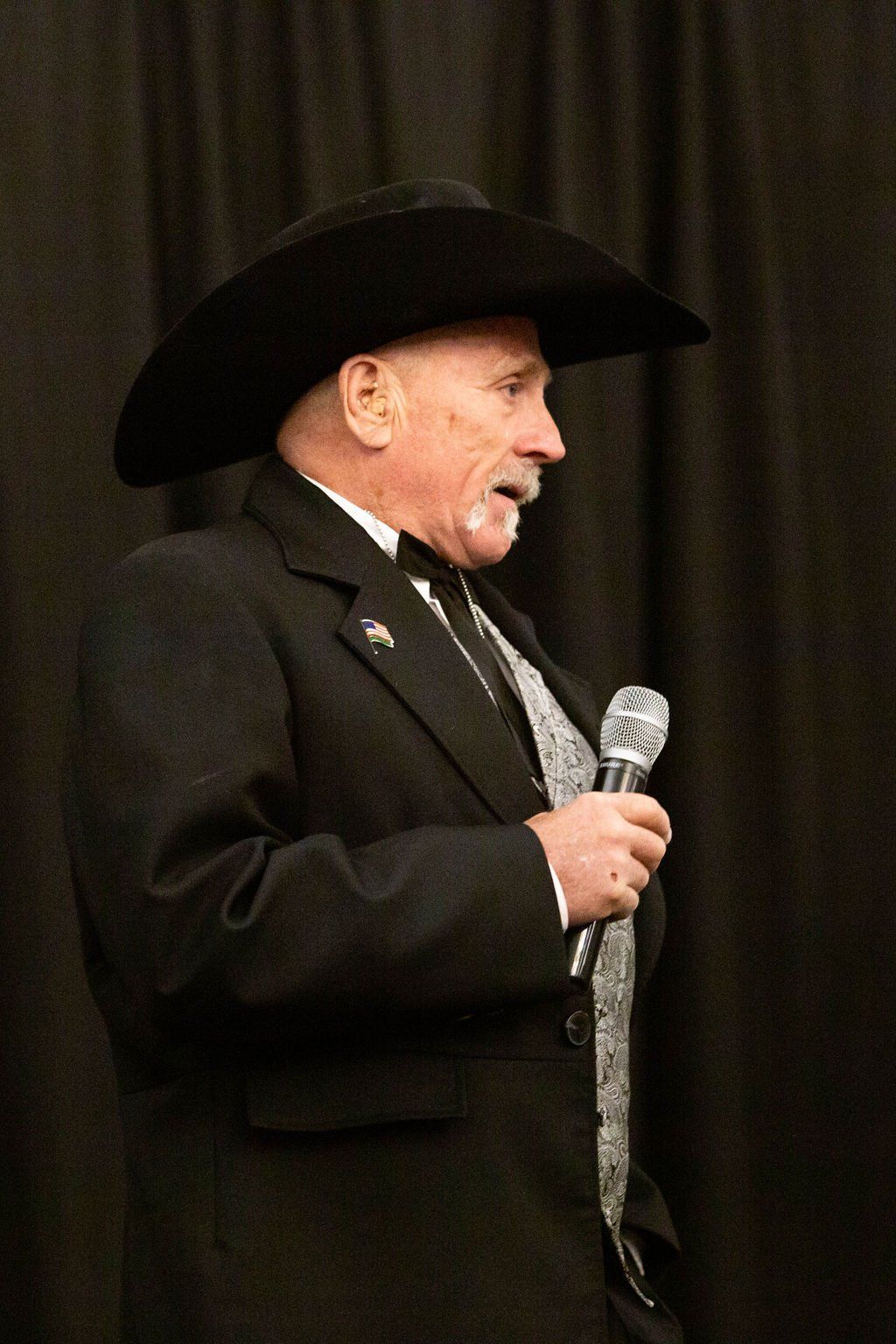 A person wearing a black cowboy hat, suit, and patterned tie holds a microphone while speaking against a black backdrop.