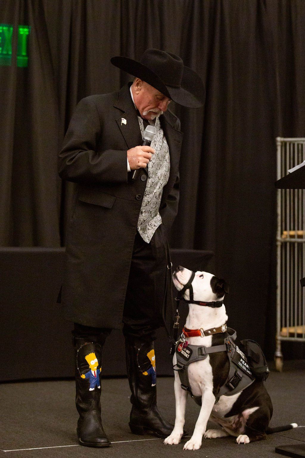 A person in a black coat, cowboy hat, and cartoon-themed boots holding a microphone, looking down at a harnessed dog.