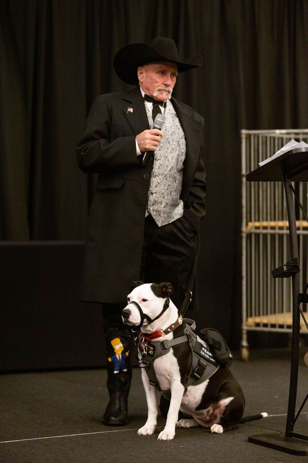 A person in a black coat, patterned vest, and cowboy hat speaks into a microphone next to a black-and-white service dog.
