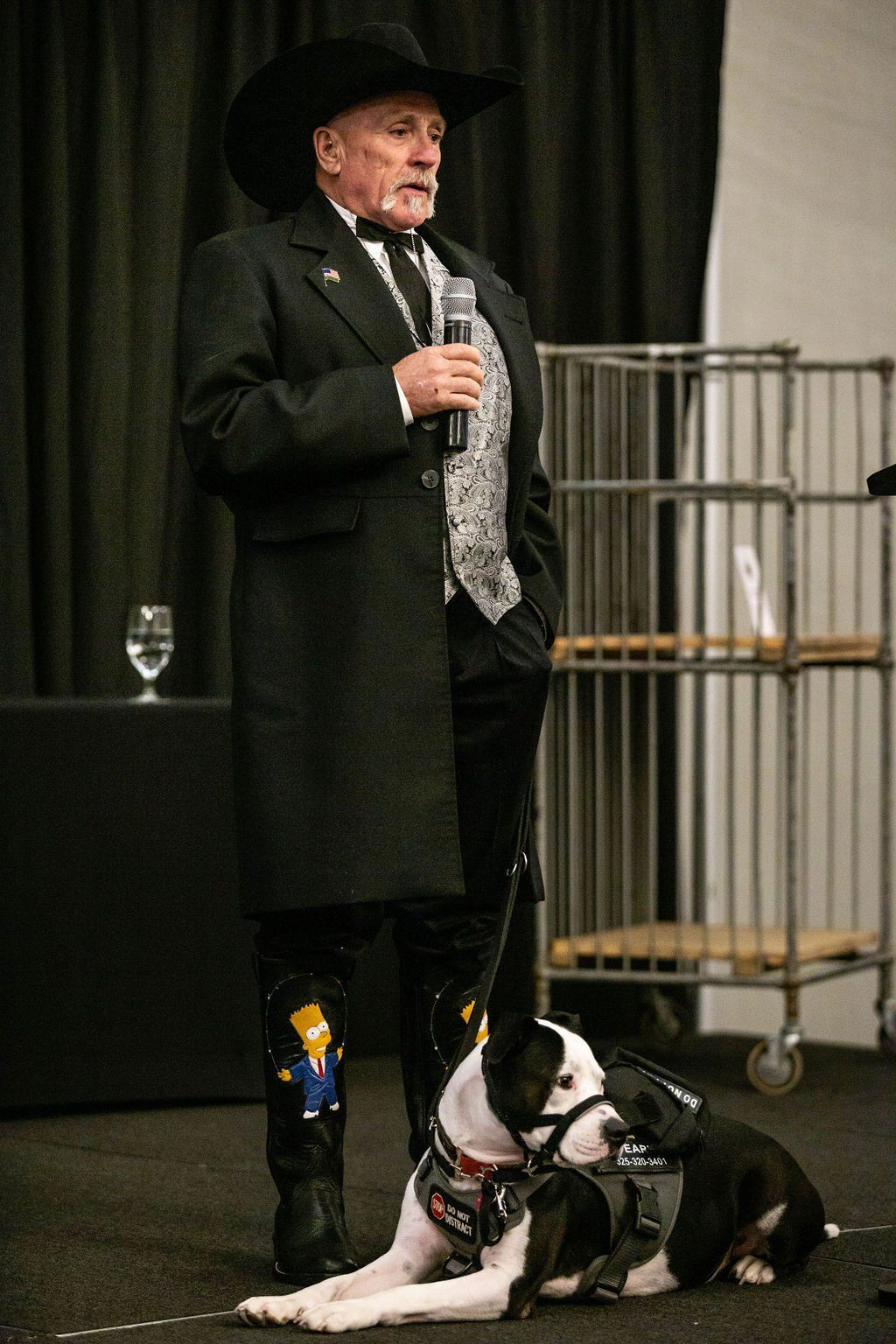 A speaker in a suit, black cowboy hat, and boots decorated with Bart Simpson sits with a service dog on a stage.