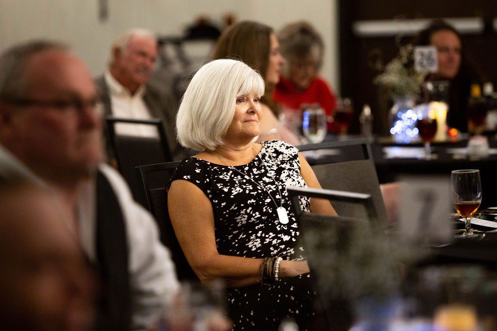 A group of people sit at tables in a dimly lit, banquet-style room, focused on a presentation or speaker.