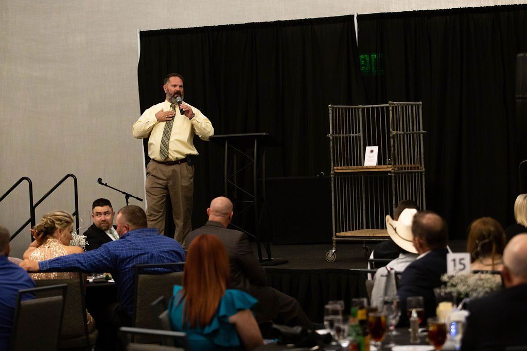 A man in a light shirt and tie speaks into a microphone at a podium during an event with a seated audience.