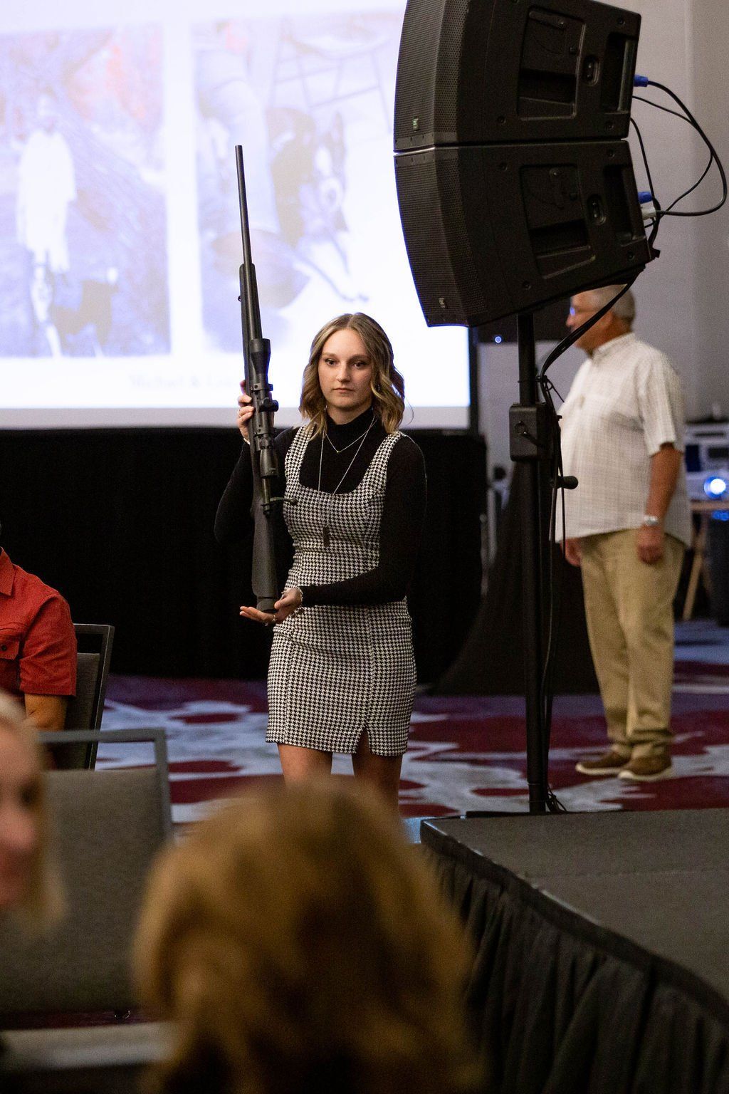 A young person in a black turtleneck and checkered dress holding a scoped rifle while standing in a conference room.