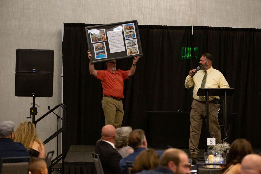 A person in an orange shirt holds up a framed collage on a stage, while a person in a tie speaks into a microphone nearby.