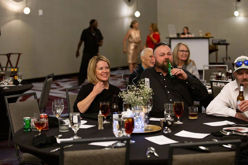 A group of people sitting at a round table with drinks in a dimly lit banquet hall, some smiling and talking.