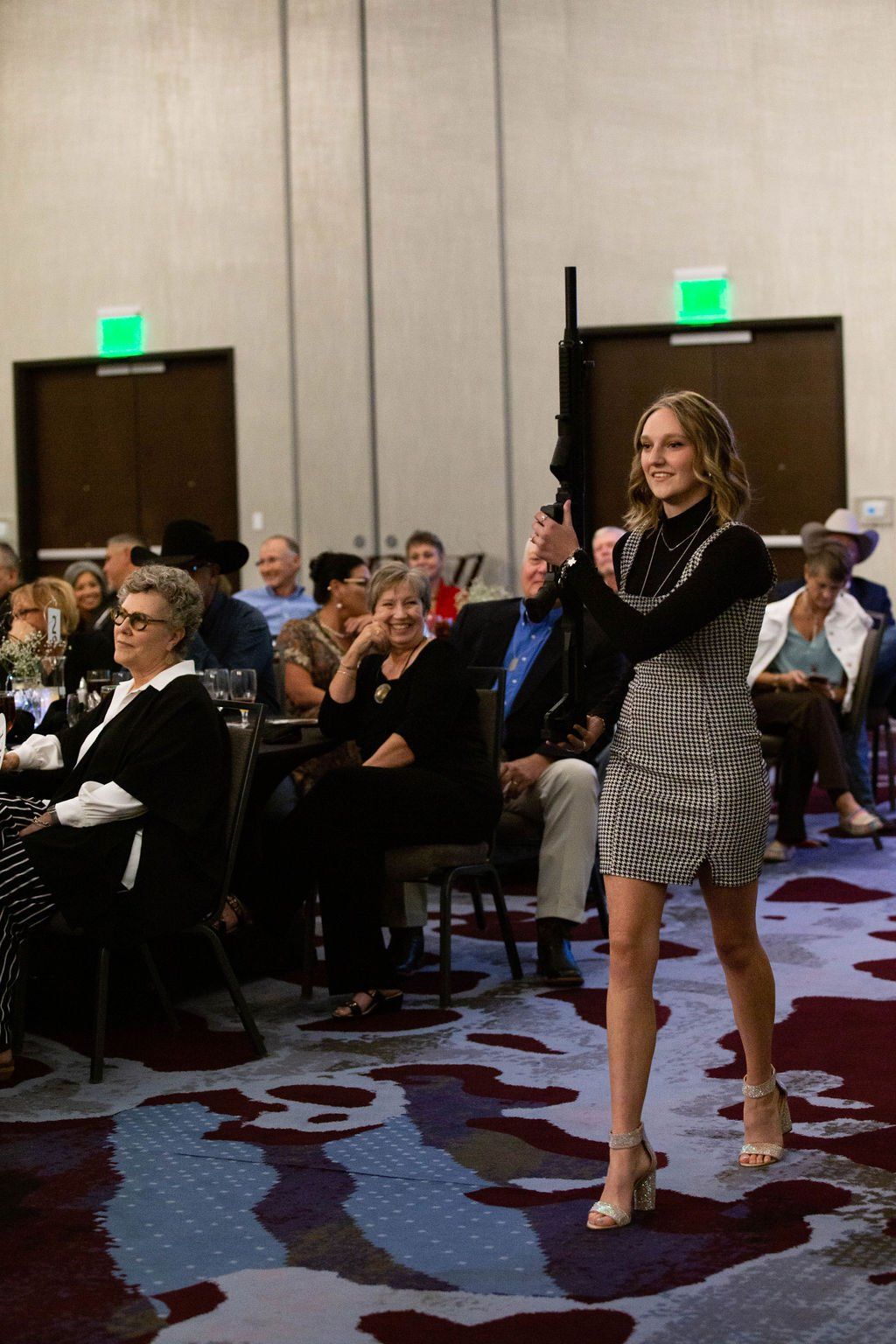 A person in a patterned dress walks across a carpeted event hall, holding a long black rifle upright in one hand.