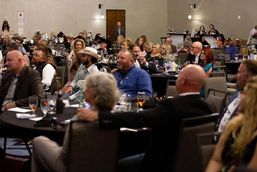 A group of people sitting at tables in a conference room, listening and looking toward the front of the hall.