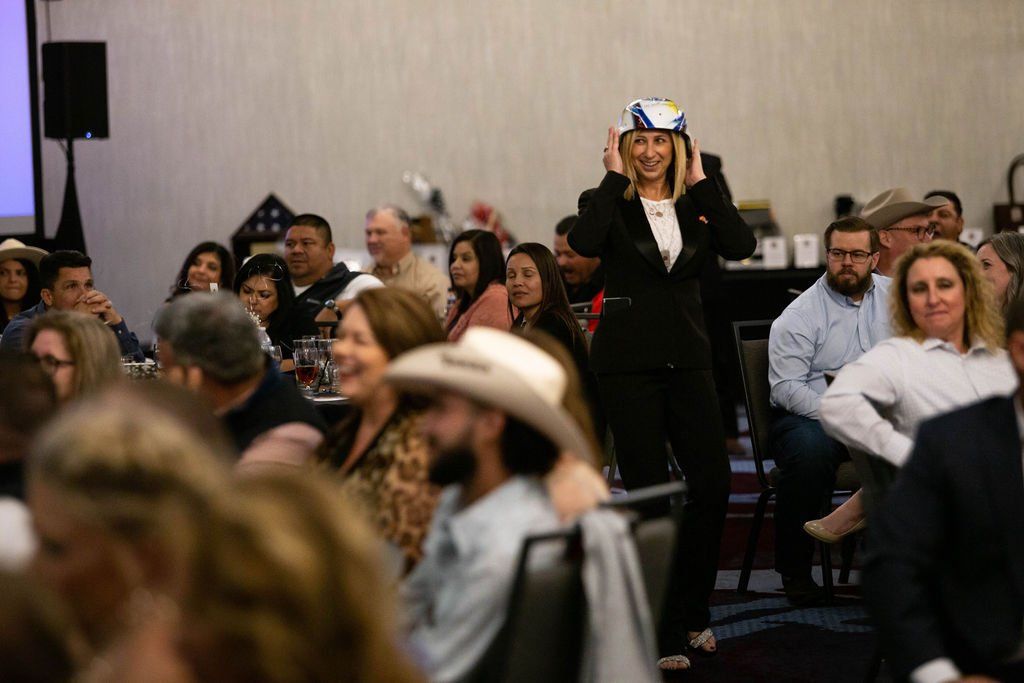 A woman in a blazer adjusts a white hard hat while standing in a crowded, dimly lit conference hall.