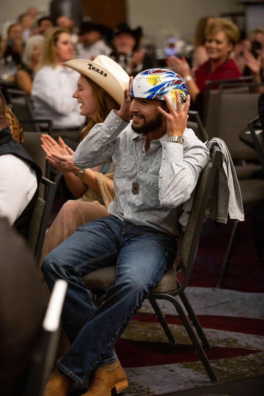 A smiling person sits in an audience, putting on a patterned protective helmet, while a person beside them applauds.