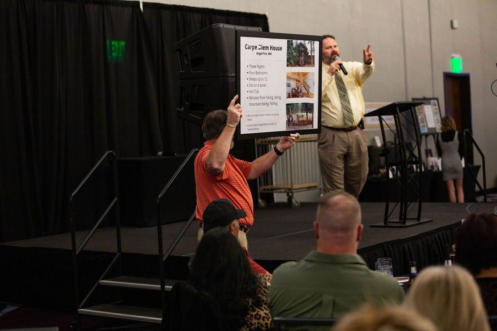 A person on a stage holds up a poster with photos and text for an audience, while a speaker gestures nearby.