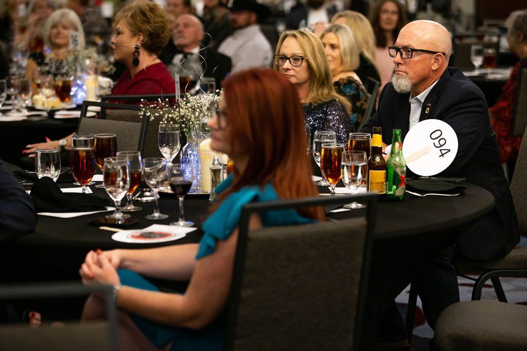 Attendees in formal attire sit at round tables in a banquet hall, listening to a speaker during a seated dinner event.