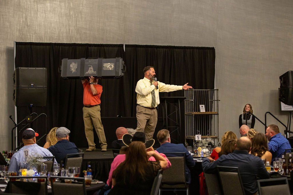 A speaker addresses an audience while a person behind them holds up a display case on a stage with black curtains.