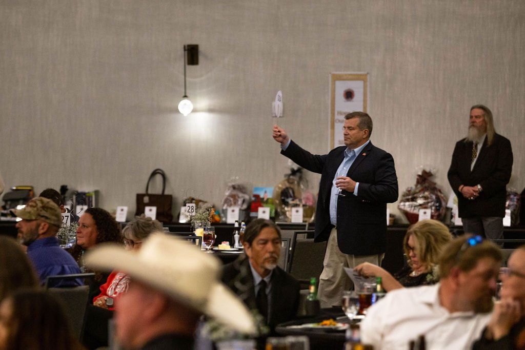 A person in a suit raises a bidding paddle at a charity auction held in a well-lit banquet hall with seated guests.