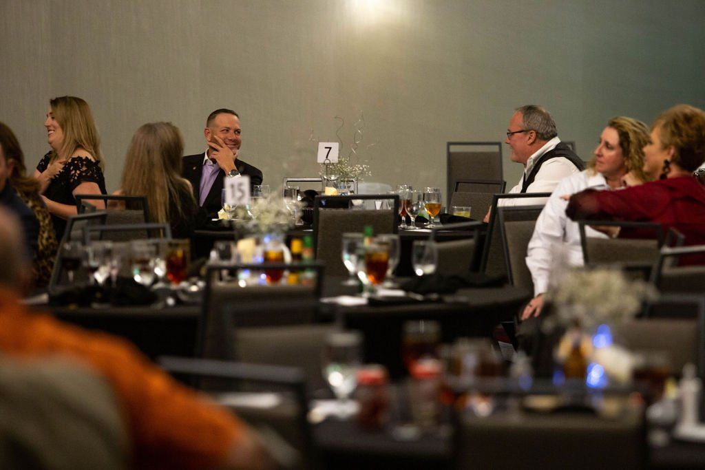 People at tables during an indoor event, talking and smiling in a dimly lit banquet hall.