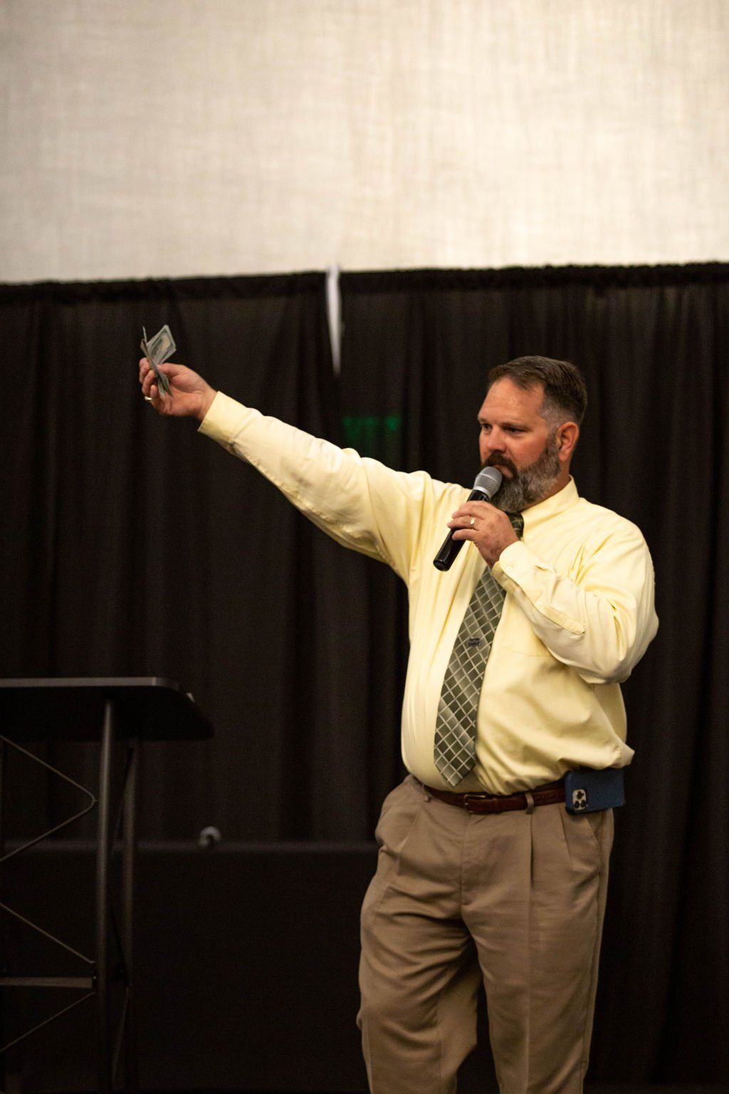 A man in a yellow shirt and tie speaks into a microphone while holding up a small object, standing before a black curtain.