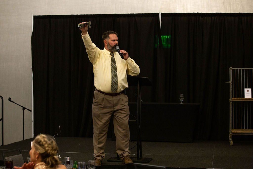 A person in a yellow shirt and tie stands on a stage with a microphone, holding an object up with their right hand.