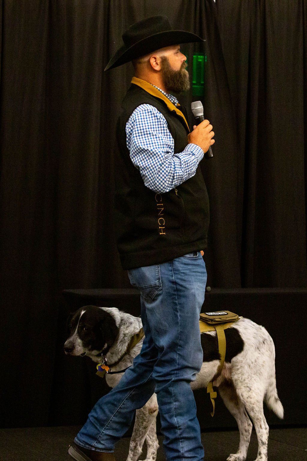A bearded man in a cowboy hat and vest stands on a stage with a microphone, accompanied by a spotted service dog.
