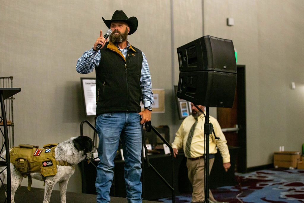 A man in a cowboy hat and vest speaks into a microphone on stage, standing beside a dog wearing a tan tactical harness.