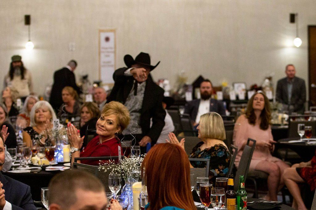 A person in a cowboy hat points toward the audience during a formal dinner event in a banquet hall.