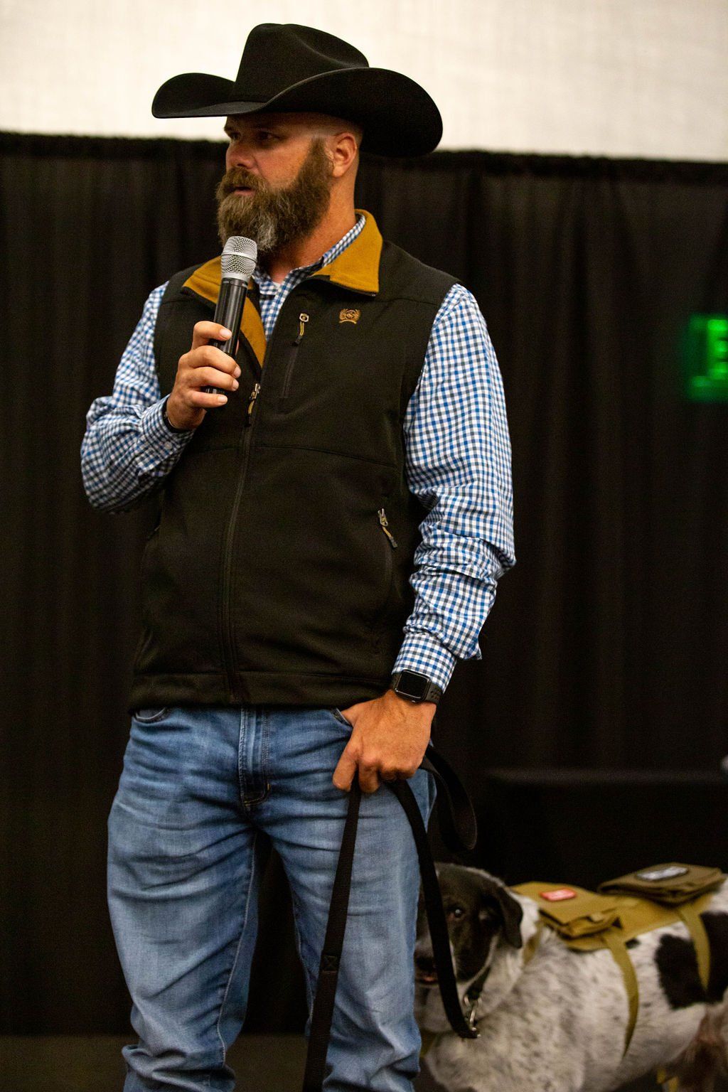 A bearded man in a cowboy hat and vest speaks into a microphone while holding the leash of a dog wearing a tactical vest.