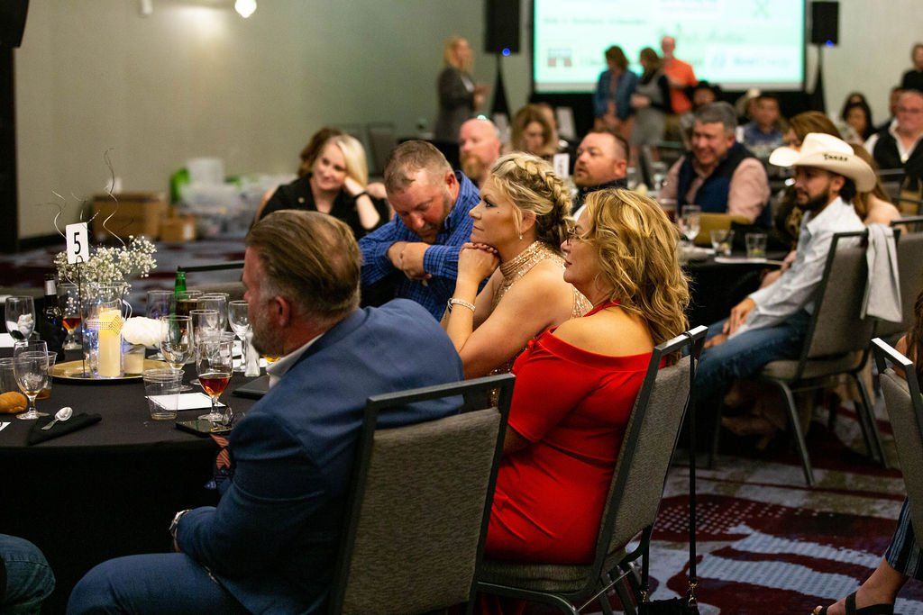 Attendees at a formal event sit at tables, looking toward a presentation screen in a dimly lit conference room.