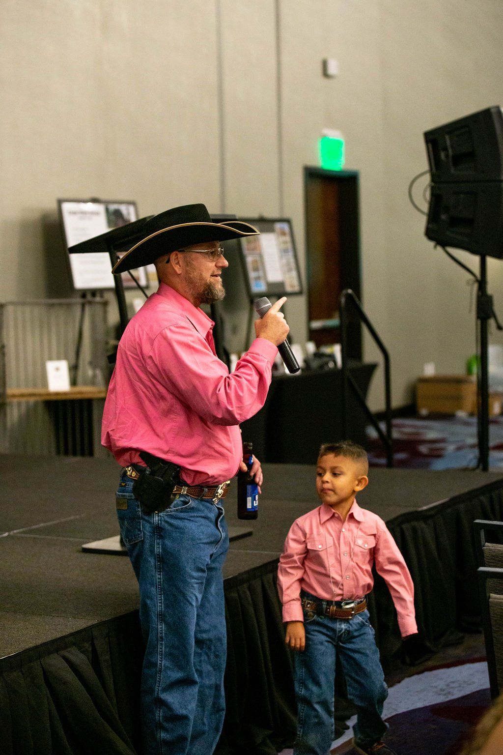 A person in a cowboy hat and pink shirt speaks into a microphone while a child stands beside them on a stage.