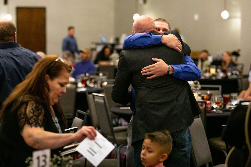 Two men in a warm embrace in a dining hall, with a woman examining a document and a child looking forward in the foreground.