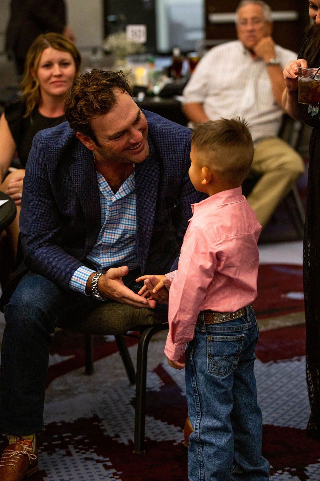 A man in a blue blazer and button-down shirt smiling and interacting with a child wearing a pink shirt and blue jeans.
