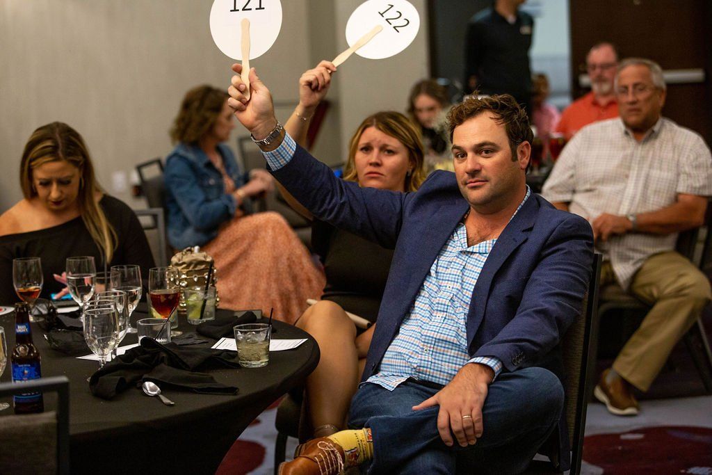 A man holds up two auction paddles at a table while others sit nearby at a banquet event.